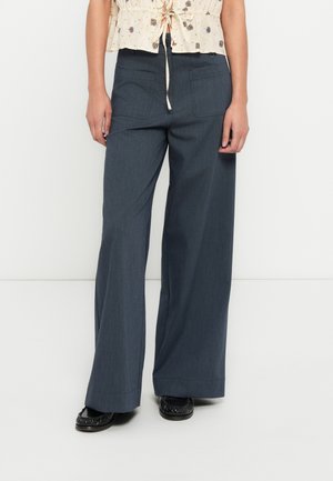 Weit geschnittene Hose aus dunkelgrauem Stoff mit strukturierter Oberfläche, ausgestattet mit zwei Gesäßtaschen und gerader Silhouette, kombiniert mit schwarzen Schuhen.