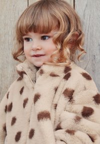 Veste en polaire beige avec des taches brunes, fermeture éclair à l'avant et col montant. Texture douce et design décontracté. Fond en surface en bois.