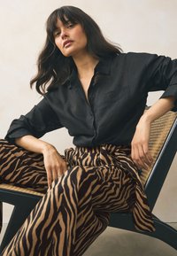 Camisa negra abotonada con tela texturizada, combinada con pantalones de pierna ancha con rayas de cebra en marrón y beige, sentado en una silla tejida.