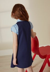 Robe bleu marine avec un corsage ajusté et une jupe évasée, associée à de courtes manches bouffantes bleu clair ; le fond présente une chaise rouge.
