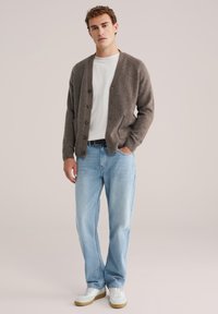 Temno rjav volnen kardigan s gumbi, oblečen čez belo majico, povezan z svetlo modrimi jeans hlačami in belimi teniskami.