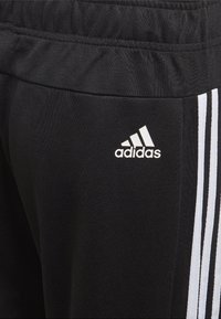 Pantalones deportivos negros hechos de una tela suave que cuentan con una cinturilla elástica, logo blanco de Adidas y tres rayas blancas a lo largo del costado.