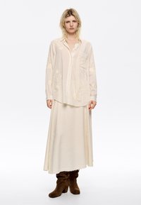Camicia color crema con ricamo floreale, dotata di tasca e dettagli in frange. Abbinata a una gonna lunga coordinata e stivali in suede marrone.