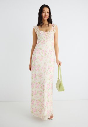 Kvinde iført en hvid maxikjole med pink og grøn blomstret print, flæse-remme, holder en lysegrøn håndtaske, stående på hvidt gulv.