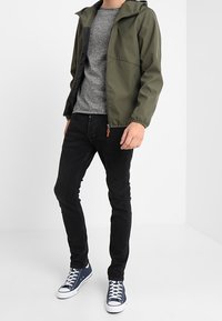 Grön lättviktsjacka med en grå tröja under, svarta jeans och blå sneakers. Jackan har en huva och dragkedjor.