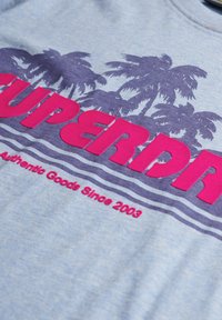 Jasnoniebieska koszulka z grafika fioletowej palmy oraz wyrazistym różowym napisem "SUPERDRY". Zawiera czerwony tekst "Autentyczne Towary Od 2003".