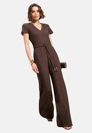 Frau trägt einen strukturierten braunen Jumpsuit mit kurzen Ärmeln, V-Ausschnitt und weiten Beinen, kombiniert mit einem goldenen Manschettenarmband und einer braunen Clutch.