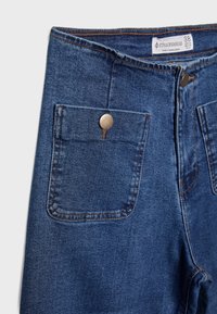Jean en denim bleu taille haute avec poches plaquées avant fermées par des boutons-pression métalliques et une étiquette de marque Stradivarius visible à l'intérieur de la ceinture.