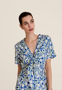 Blouse à manches courtes avec un nœud sur le devant, comportant un motif abstrait bleu, vert et blanc sur un tissu clair.