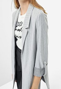Vrouw in een lichtgrijze gestreepte blazer met opgerolde mouwen over een wit T-shirt met opdruk en zwarte broek.