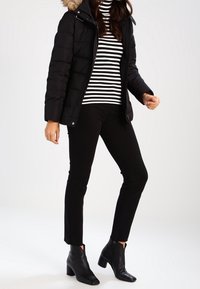 Musta toppatakki, jossa on turkisvuorattu huppu, yhdistettynä mustavalkoraidalliseen poolopaitaan ja mustiin istuviin housuihin. Mustat nilkkurit näkyvissä.