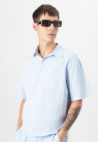Hellblaues Hemd mit kurzen Ärmeln aus strukturiertem Stoff, Knopfleiste vorne und einer einzelnen Brusttasche. Schwarze, rechteckige Sonnenbrille rundet den Look ab.