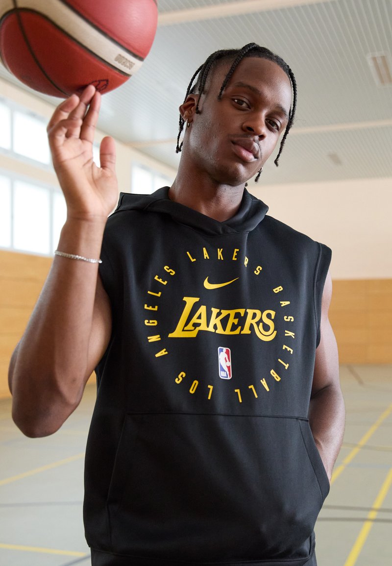 Melnā bezpiegu vestē ar dzelteno "Lakers" logo un NBA emblēmu, kuru valkā cilvēks, kurš telpās pagriež basketbola bumbu uz viena pirksta.
