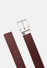 Brązowy skórzany pasek o teksturze z metalową klamrą w kolorze srebrnym, pokazujący koniec z otworami do regulacji i detalem zapięcia na białym tle.