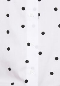 Chemise blanche à pois noirs, fermeture à boutons et texture de tissu lisse avec une coupe classique.