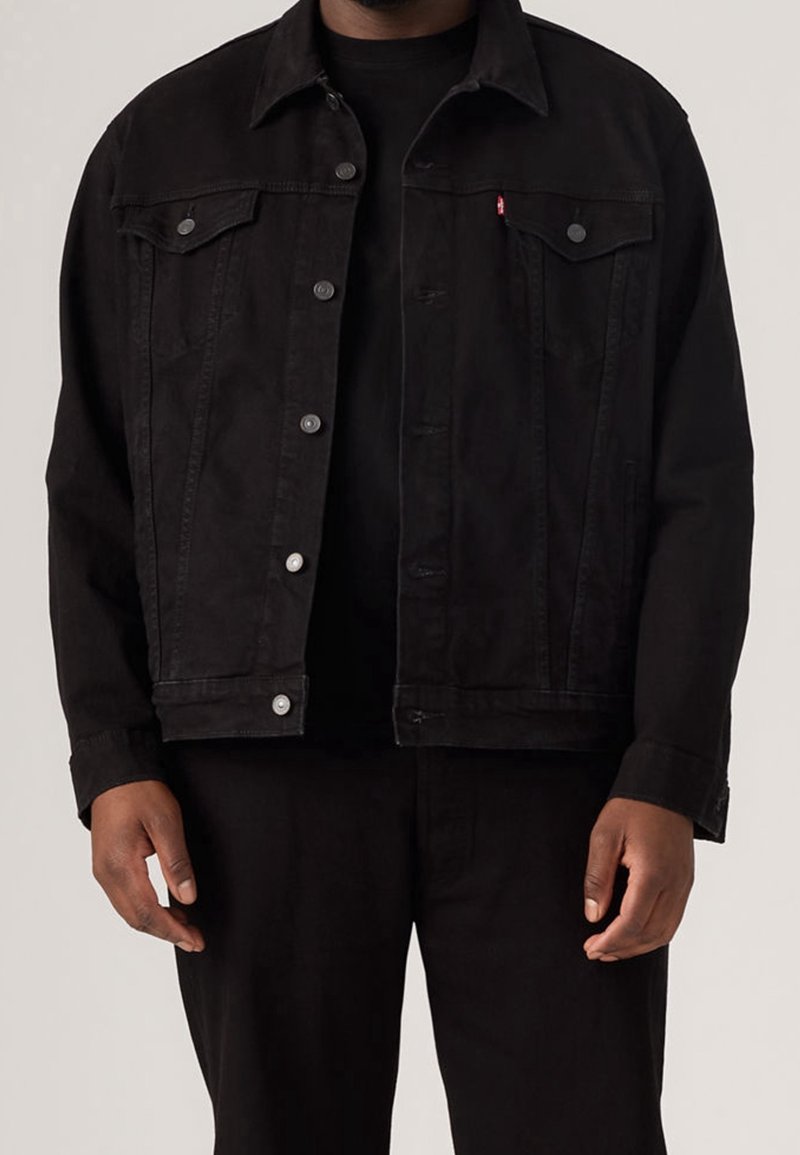 Homme portant une veste en jean noire par-dessus une chemise noire et un pantalon noir, debout devant un fond clair uni.