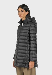 Black puffer jacket with horizontal quilting, high collar, front zipper, and hood. Smooth, shiny material; logo patch near the hem.