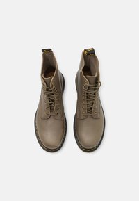 Botas de tobillo de cuero marrón con cordones, costuras de acento amarillas y una lengüeta en el talón. Textura suave con punta redondeada y suela resistente.