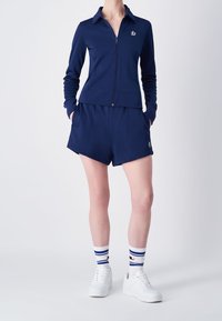 Giacca sportiva blu navy con zip e colletto, abbinata a pantaloni corti coordinati. Il modello indossa sneakers bianche e calze a righe.