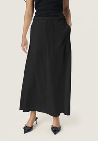 Zwarte maxi-rok met een platte taille en verstelbare strikjes, voorzien van zijzakken en een gladde textuur. Gepaard met zwarte puntige hakken.