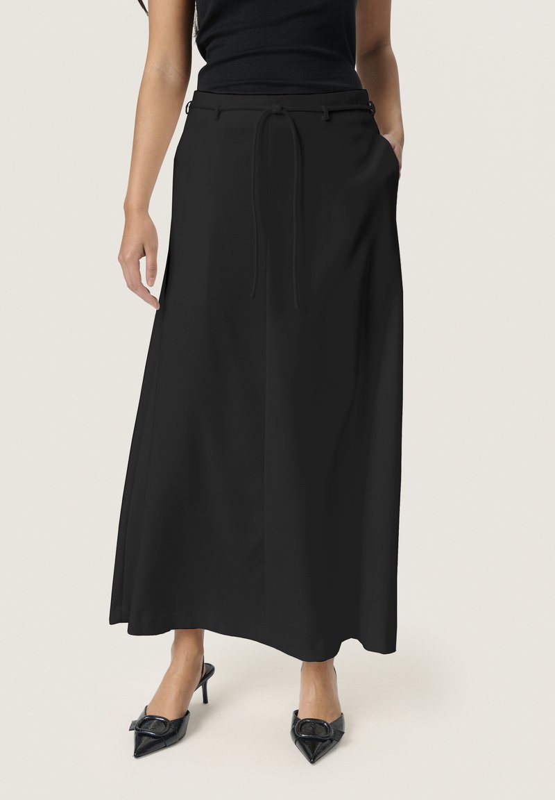 Falda maxi negra con cintura plana y lazos ajustables, con bolsillos laterales y una textura suave. Combinada con tacones negros de punta afilada.