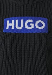 Schwarzer gestrickter Pullover mit einem auffälligen blauen rechteckigen Patch, auf dem das Wort "HUGO" in großen weißen Buchstaben steht. Die Textur ist gerippt.