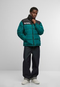 Kurtka puchowa w kolorze morskim z czarnym kołnierzem; posiada poziome pikowania i biały napis "KARL". Połączona z ciemnymi dżinsami i białymi trampkami.