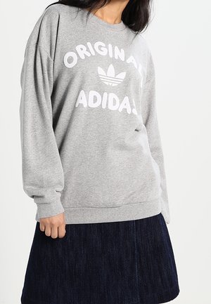Osoba nosící šedou mikinu Adidas Originals s logem a nápisem, spojenou s tmavou plisovanou sukní na jednoduchém pozadí.