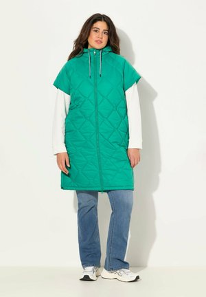 Chaleco acolchado verde con cuello alto, cierre de cremallera y mangas cortas, superpuesto sobre una camiseta blanca de manga larga, combinado con unos jeans azules.
