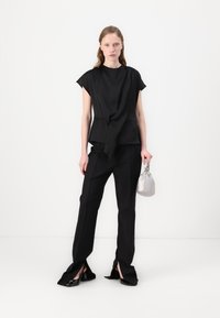 Top nero con dettagli drappeggiati, abbinato a pantaloni neri su misura con spacchetti laterali. La modella tiene in mano una piccola borsa metallica argentea.