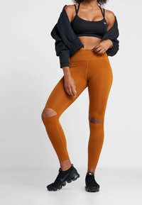 Leggings atletici in arancione ruggine con dettagli ritagliati alle ginocchia, abbinati a un reggiseno sportivo nero e a delle sneakers nere. Tessuto liscio e aderente.