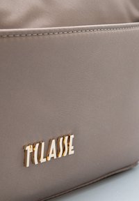 Primo piano di una borsa in tessuto taupe con logo in lettere dorate "1a CLASSE" e cuciture visibili vicino al bordo superiore.