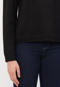 Zwarte gebreide trui met een geribde onderrand en manchetten, gecombineerd met donkerblauwe jeans. De trui heeft een gestructureerde en zachte stof.