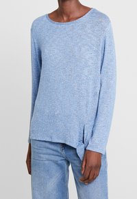 Ljusblå stickad långärmad topp med rund halsringning, sidofickor och en knuten detalj vid nederkanten. Texturerat material ger visuell variation.