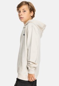 Beige hoodie met een kangoeroezak, ribgebreide mouwen en capuchon. Voorzien van een klein marineblauw logo op de borst en een zachte, gestructureerde stof.