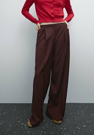 Persoon draagt losse, wijd uitlopende donkerbruine broek, rode cropped cardigan, staand op een donkere gevlekte vloer tegen een lichtgrijze muur.