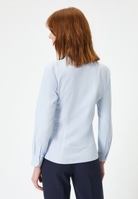 Ljusblå, långärmad blus med en figurnära design, mjukt tyg och knappade manschetter. Baksidan har subtila sömdetaljer.