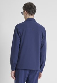 Chaqueta azul marino de tejido suave con cuello estructurado, mangas largas, puños abotonados y un pequeño detalle de etiqueta en la parte superior de la espalda.