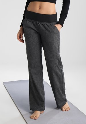 Žena stojící bosá na šedé jógové podložce, oblečená v tmavě šedých širokých kalhotách s černým pasem a krátkém černém dlouhém rukávu.