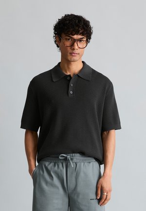 Jeune homme aux cheveux bouclés et portant des lunettes, vêtu d'un polo en maille noire et d'un pantalon gris à cordon de serrage, debout devant un fond uni.