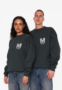 Tmavé šedé mikiny so štruktúrovaným bielym logom "M" na hrudi. Majú voľný strih, rebrované manžety a uvoľnený výstrih. Viditeľné džínsy.