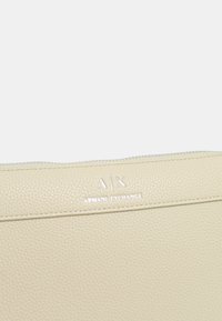Borsa a pochette in pelle beige con superficie testurizzata. Presenta il logo "A|X" e "ARMANI EXCHANGE" in argento sulla parte anteriore e una chiusura a zip nella parte superiore.
