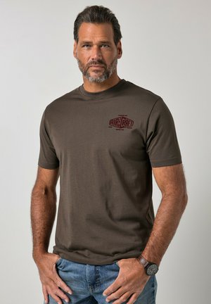 Mężczyzna z krótkimi ciemnymi włosami i brodą, ubrany w brązowy t-shirt "Aircraft", niebieskie dżinsy i zegarek na rękę, stoi z rękami blisko kieszeni.