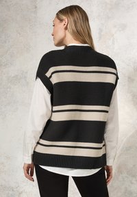 Ärmelloser, schwarzer Strickpullover mit breiten cremefarbenen Streifen. Gerippter Ausschnitt und Saum. Über einem weißen Hemd mit Kragen getragen.