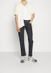 Ljust gul kortärmad skjorta med knappslå och strukturerad design, vit linne, mörkblå jeans och flerfärgade sneakers.