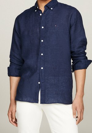 Man draagt een marineblauw overhemd met lange mouwen met opgerolde manchetten en een witte broek, staand tegen een neutrale achtergrond.