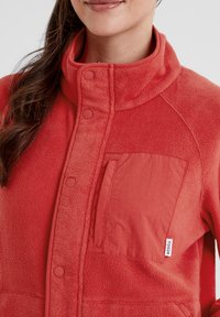 Red fleece jacket with a high collar, featuring snap buttons and a patch pocket made from a smooth fabric. Logo tag visible on side.