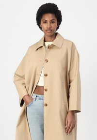 Trench coat beige, leggero, con ampio colletto e grandi bottoni marroni. Presenta tasche laterali e una vestibilità over-size. Texture liscia.