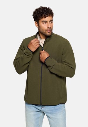 Chaqueta verde oliva con cremallera y cuello alto, textura acanalada, acentos en azul oscuro contrastantes en los puños y la cremallera. Combinada con vaqueros azul claro.