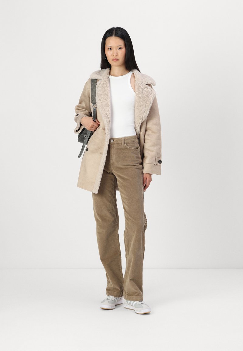 Manteau en fausse fourrure crème avec un col, porté sur un débardeur blanc et un pantalon en velours côtelé marron. Sac à bandoulière gris et baskets blanches.